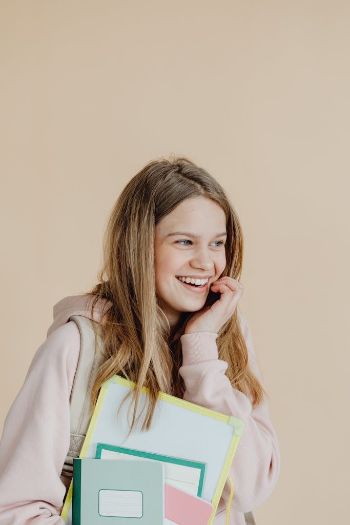 Smiling teenage girl holding notebooks against a beige background, conveying joy and readiness for school.