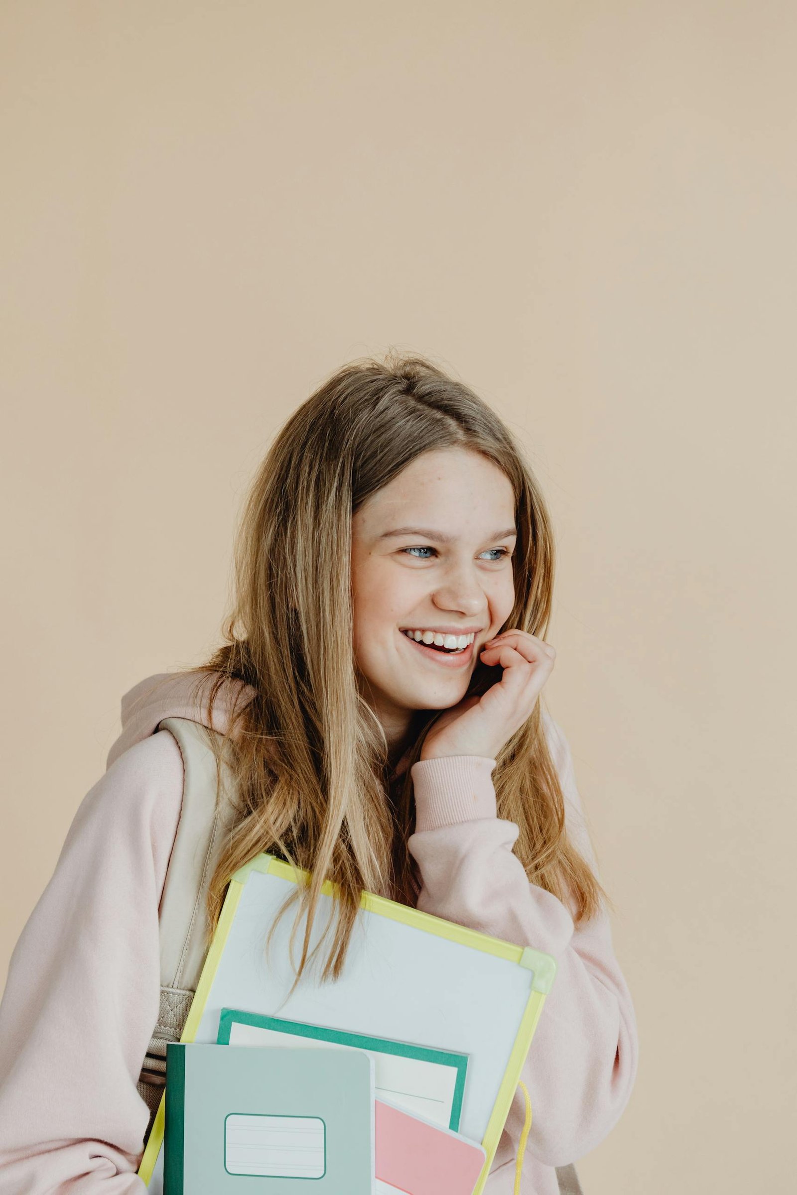 Smiling teenage girl holding notebooks against a beige background, conveying joy and readiness for school.